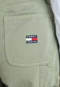 Zelená bavlněná tkanina s zadní kapsou, na které se nachází nášivka s logem "TOMMY JEANS". Čisté švy a zaoblený tvar kapsy.
