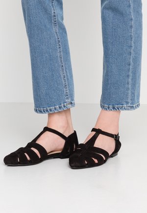 Ballerines en suede noir avec un design ouvert, dotées de plusieurs lanières et d'une boucle. La semelle est plate, complétée par une légère texture.