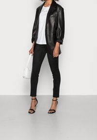 Blazer oversized de cuero negro, camiseta blanca con texto, pantalones skinny de ante negro y tacones negros con tiras. Sostiene un bolso blanco.