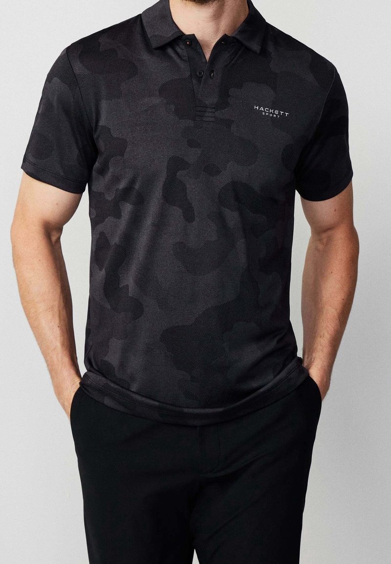 Homme portant un polo noir à manches courtes avec un motif camouflage et le logo "Hackett Sport" sur la poitrine, mains dans les poches d'un pantalon noir.