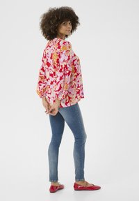 Gėlių bliuska rožinės ir oranžinės spalvų atspalviuose, su ilgomis puffed rankovėmis; derinama su mėlynomis siaurėjančiomis džinsomis ir raudonais butais.