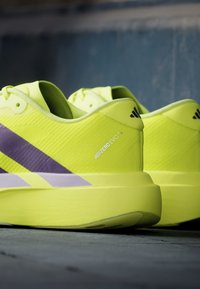 Φωτεινά κίτρινα παπούτσια τρεξίματος Adidas Adizero Evo SL με μωβ ρίγες τοποθετημένα σε γκρι επιφάνεια με σκουρό μπλε φόντο.