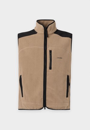Homme debout portant un gilet beige sans manches sur un t-shirt blanc, un pantalon noir ample et des chaussures noires sur un fond clair uni.