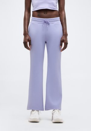 Άτομο που φοράει ανοιχτό μωβ καμπάνα φόρμα με κορδόνι στη μέση, λευκά αθλητικά παπούτσια και κοντό τοπ, στέκεται μπροστά σε απλό φόντο.