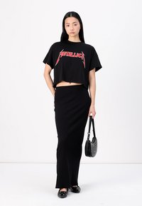 Černé zkrácené tričko s červenou grafikou "Metallica", spárované s černou přiléhavou maxi sukni. Nese malou černou tašku a má na sobě ploché boty.