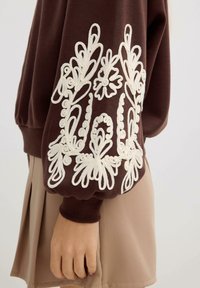 Sudadera marrón con un patrón floral blanco bordado en la manga. El diseño presenta espirales y bucles. Acompañada de una falda beige.