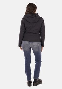 Giacca nera con cappuccio, finitura liscia e opaca, design slim, abbinata a jeans blu e stivali neri chunky, vista da dietro.