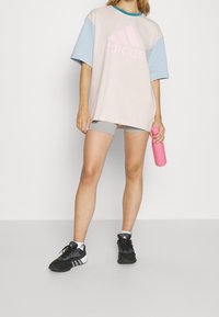 Ljust rosa oversized t-shirt med blå ärmar och en stor rosa Adidas-logotyp. Gråa träningsshorts och svarta sneakers med vita accenter.