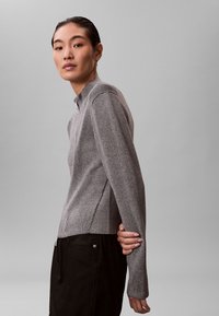 Szary sweter z prążkowanej dzianiny z wysokim kołnierzem, bocznymi rozcięciami i długimi rękawami. W połączeniu z ciemnymi spodniami, wyróżnia się charakterystyczną fakturą i nowoczesną sylwetką.