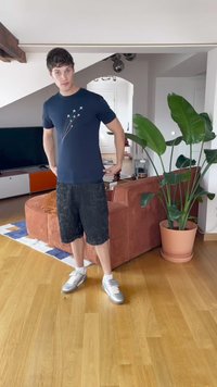 Tmavomodré tričko s grafickým motívom lietadla, v kombinácii s čiernymi rifľovými šortkami. Stojí na drevenej podlahe pri hnedej pohovke a kvetináči.