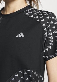 Detailní záběr na černé atletické tričko s krátkým rukávem, s bílým trojúhelníkovým vzorem a malým bílým logem na hrudi.