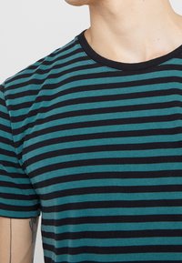 Persona vestida con una camiseta de mangas cortas a rayas horizontales en color teal y negro, que muestra el cuello, el hombro y un tatuaje parcial en el brazo izquierdo.