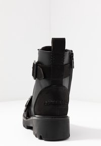 Czarny botek na kostkę wykonany ze skóry i zamszu, wyposażony w zamek błyskawiczny, pasek z klamrą oraz teksturowaną gumową podeszwę.
