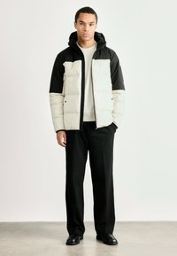 Puffjakke i creme og sort med frontlynlås, hætte og quiltet tekstur. Parret med sorte bukser og sko. Minimalistisk design.