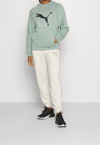 Mint green hoodie with a black graphic design, paired with cream joggers featuring a gold logo. Black athletic shoes complete the outfit.