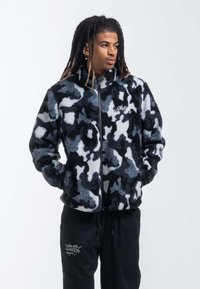 Fleecepolo med dragkedja som har ett blått, grått och svart kamouflage-mönster. Den har en hög krage och framdragkedja med broderad logotypdetalj.