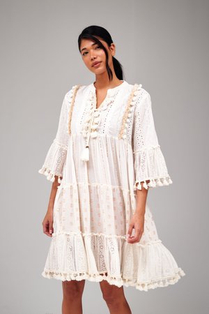 Femme portant une robe blanche mi-longue en dentelle avec des pampilles et des manches évasées trois-quarts, debout devant un fond gris uni.