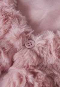 Rosa faux fur-tyg med en mjuk, fluffig textur. Inkluderar en rund, ljusrosa knappdetalj, placerad mot en slät rosa bakgrund.