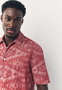 Camicia a maniche corte rossa con motivo geometrico, realizzata in tessuto testurizzato. Presenta chiusura con bottoni e colletto appuntito.