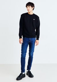 Sweatshirt noir à col rond, jeans en denim et baskets noires. Design simple avec des poignets et un ourlet côtelés. Le modèle se tient devant un fond neutre.