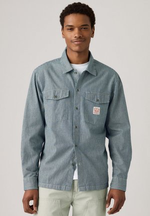 Blue and white striped button-up shirt with two chest pockets, a small logo patch, and a collared neckline. Cotton material with a textured finish.