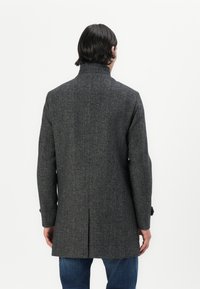 Manteau en mélange de laine gris avec col montant, design ajusté, fente au dos et poignets boutonnés, porté sur un jean bleu.