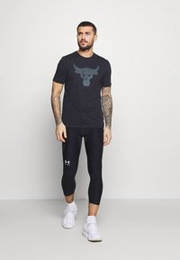 Muž s tetovaniami v čiernom tričku s logom čierneho býka, čiernych legínach do polovice lýtok a bielych teniskách kráča pred jednofarebným sivým pozadím.