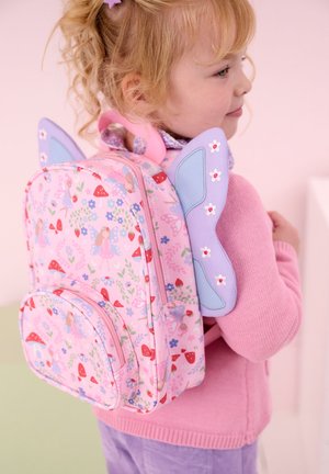 Junges Kind trägt einen rosa Pullover und lila Hosen und trägt einen rosa Rucksack im Feen-Thema mit Schmetterlingsflügeln und Blumenmustern.