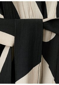 Černý a béžový látkový pásek uvázaný kolem oděvu s výrazným abstraktním geometrickým vzorem a texturovaným materiálem.