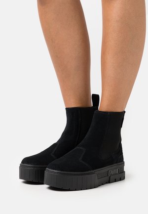 Bottes chevilles en daim noir avec semelles plateforme épaisses en caoutchouc et panneaux latéraux élastiques portées sur des jambes nues sur fond blanc.