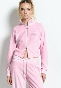 Młoda kobieta z wysokim kucykiem, ubrana w jasnoróżową kurtkę sportową na zamek i dopasowane spodnie, stojąca na białym tle.