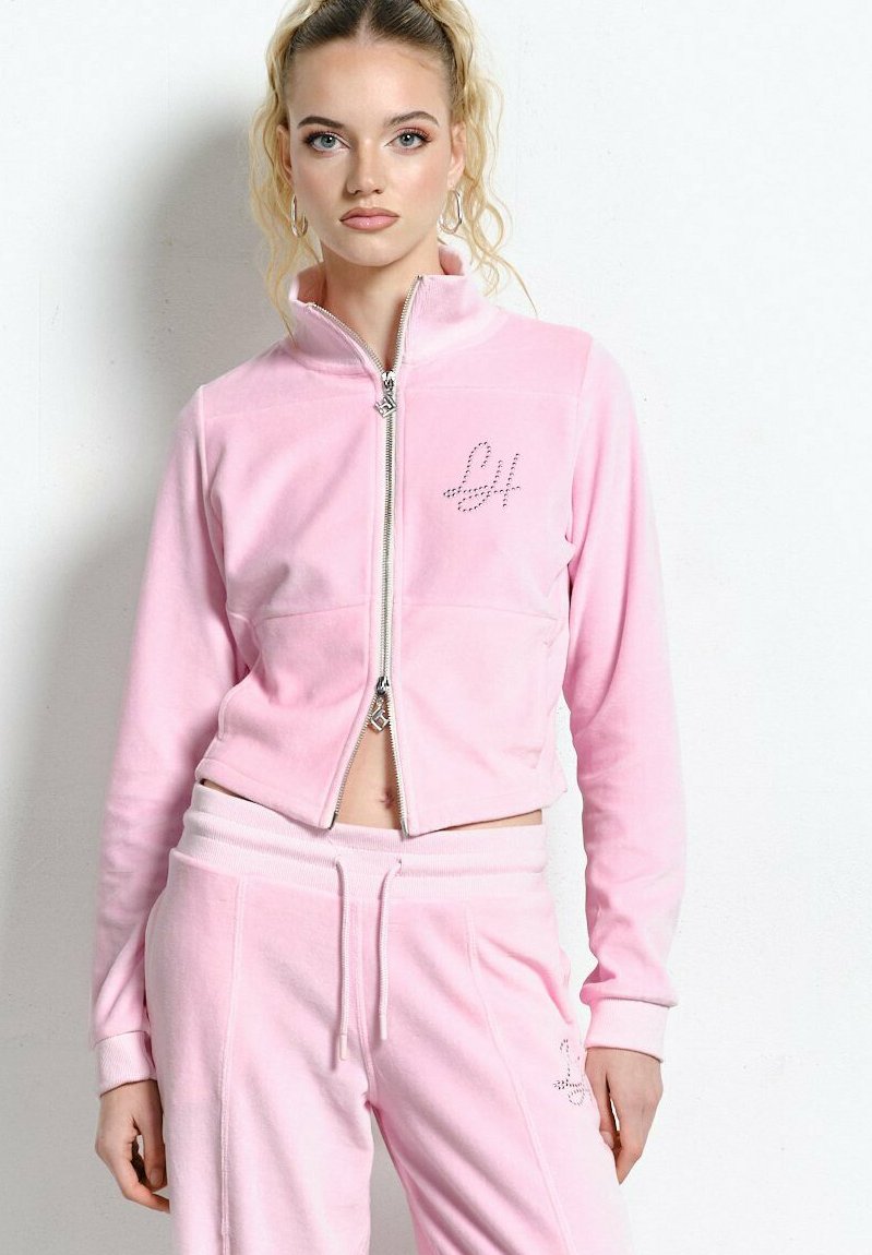 Młoda kobieta z wysokim kucykiem, ubrana w jasnoróżową kurtkę sportową na zamek i dopasowane spodnie, stojąca na białym tle.