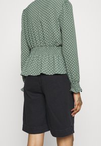 Blusa verde con pois, con vita arricciata e maniche lunghe, abbinata a pantaloni shorts in denim scuro, caratterizzati da una vestibilità comoda e orli risvoltati.