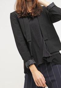 Blazer nero con un design strutturato, dotato di maniche lunghe, apertura frontale e una maglietta nera con texture sottostante, indossato con pantaloni a righe.