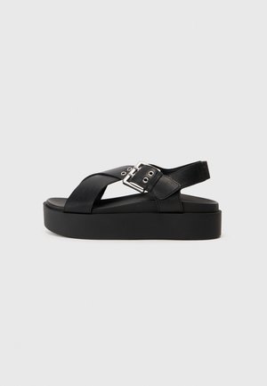Black platform sandals with cross straps, made of smooth synthetic material. Features a wide buckle and flat sole for comfort.