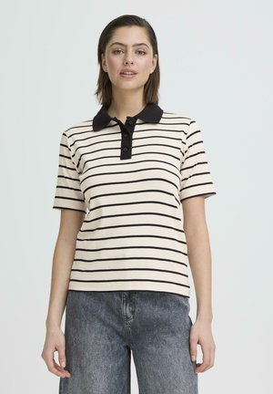 Femme aux cheveux bruns courts portant un polo crème à manches courtes avec des rayures horizontales noires et un jean gris foncé, debout devant un fond gris.