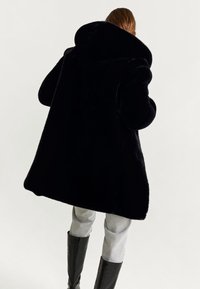 Zwarte faux fur jas met een brede kraag, lange mouwen en een losse pasvorm. Soepele textuur, reikt tot halverwege de dij, gecombineerd met grijze broek en hoge zwarte laarzen.