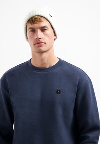 Marineblauwe sweatshirt van textuurstof, met ribbelkraag en zwart hexagonaal logo-embleem. Gecombineerd met een lichtgrijze ribbelbeanie.