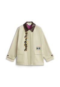 Beige jas met een donkerbruine leren kraag, een voorkant opgestikte zak met een bloemenlabel en verticale bloemrijke borduursels aan de linkerkant.