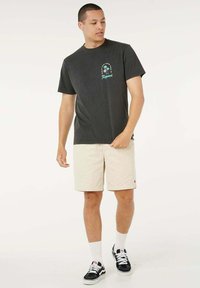 T-shirt in cotone grigio scuro con stampa grafica e testo, abbinata a pantaloncini beige chiaro. Scarpe da ginnastica nere e calzini bianchi completano il look.