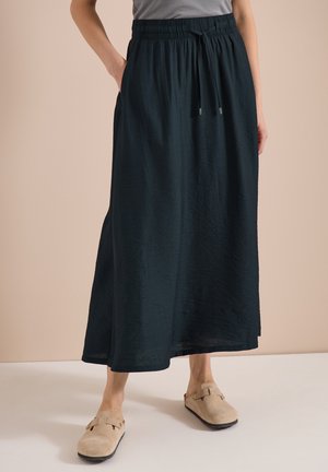 Femme debout, les mains dans les poches, portant une longue jupe noire avec taille à cordon et des chaussures beige à enfiler sur fond beige.