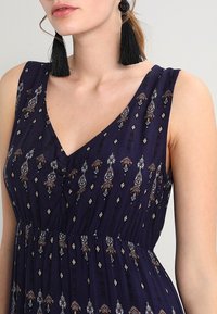 Vrouw in een mouwloos marineblauw jurkje met een V-hals en ingewikkelde geometrische patronen, gecombineerd met lange zwarte franjenoorbellen.