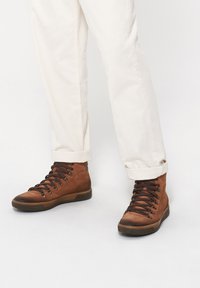 Brązowe skórzane buty typu high-top z okrągłym noskiem, z metalowymi oczkami, czarnymi sznurówkami i teksturowaną gumową podeszwą. Połączone z białymi spodniami.