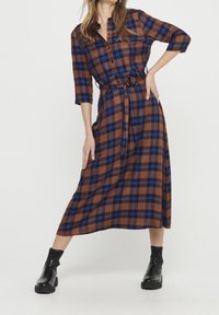 Hnědé kostkované šaty vyrobené z lehkého materiálu, s tříčtvrtečními rukávy, páskem v oblasti pasu a střední délkou. Černé kotníkové boty doplňují celý outfit.