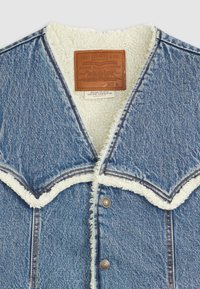 Denimjakke med en blå stofoverflade, hvid fleeceforing og en broderet krave. Har et læder mærketag og metalknapper.