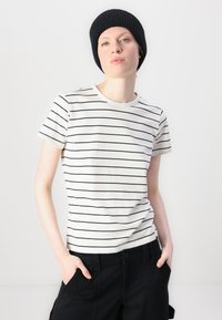 T-shirt in cotone a righe bianche con sottili righe orizzontali nere. Collo a giro, maniche corte, abbinato a pantaloni neri, indossato in modo informale.