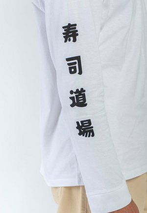 Chemise blanche à manches longues avec des caractères japonais verticaux noirs sur la manche portée par une personne en pantalon beige.