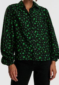 Camicia nera con piccolo motivo floreale verde, dotata di una chiusura frontale con bottoni, colletto e maniche a sbuffo raccolte ai polsini.
