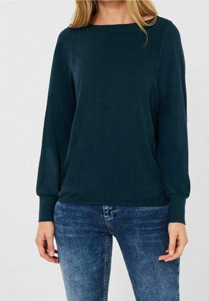 Femme portant un chemisier vert foncé à manches longues avec ourlet élastique et un jean bleu sur un fond uni clair.