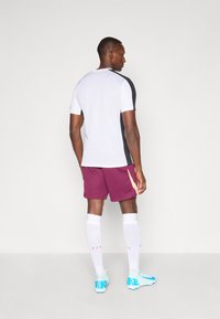Sportovní outfit skládající se z bílého a černého trička s krátkým rukávem, vínových šortkách, bílých ponožkách a světle modrých fotbalových kopačkách.